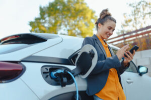 Woman checking her phone while leaning against her EV charger and using an electric vehicle charger installed in her home.