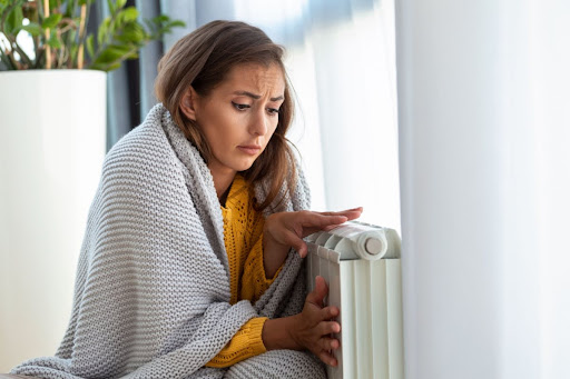 A woman looking cold with a blanket wrapped around her as she puts her hands on a radiator in a home.