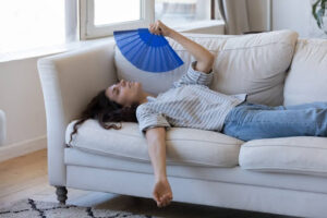 Woman splayed out on couch fanning herself with a hand fan because of broken air conditioner.