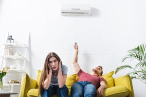 Woman sweating on the couch as her man holds up remote to broken AC unit.