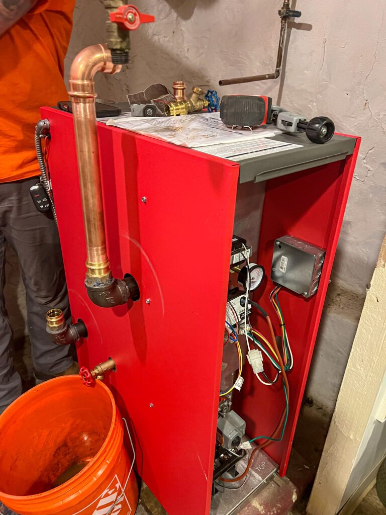 New red boiler installation in progress with exposed copper piping, electrical wiring, and tools on top, prepared for residential heating system upgrade.
