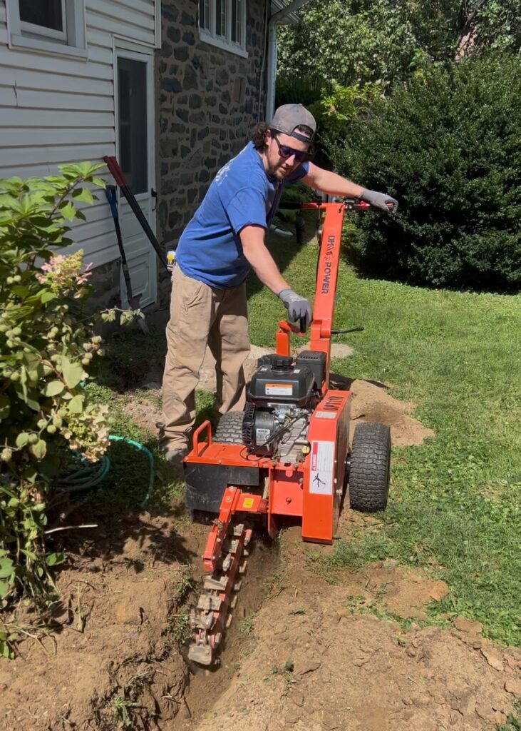 Electrician using a trencher to dig for a detached garage subpanel installation in Philadelphia backyard.