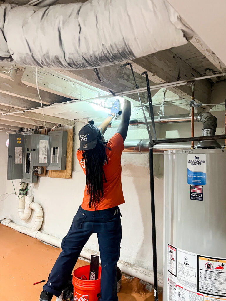GEN3 HVAC technician working in a basement, wiring above ductwork near the water heater and electrical panels.
