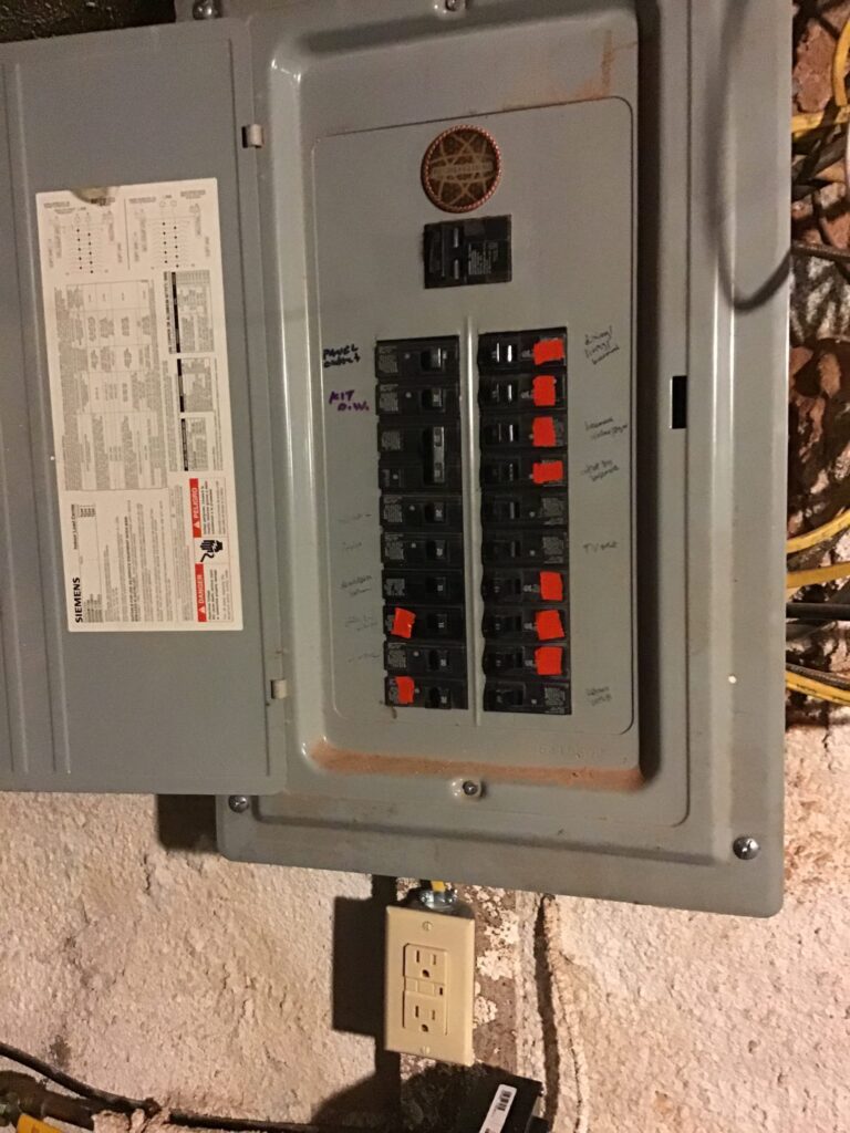 Residential electrical breaker panel with handwritten circuit labels and GFCI outlet below, shown before an electrical upgrade.